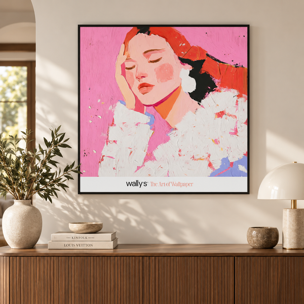 A framed art print featuring a stylized portrait of a woman with red hair and a white textured coat against a pink background, hanging on a beige wall above a wooden console table with books, a vase, and a lamp.