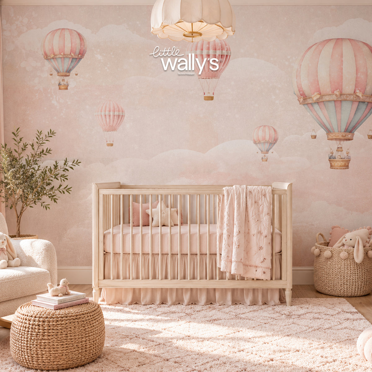 A styled nursery room featuring a light wood crib with a pink and white star-patterned blanket, set against a wall with pink and blue hot air balloon wallpaper. A white armchair, a woven ottoman, and a basket with plush toys are arranged in the room.