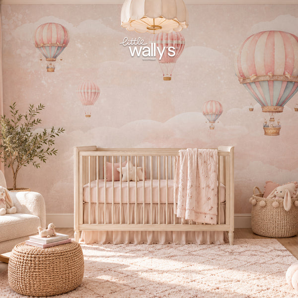 A styled nursery room featuring a light wood crib with a pink and white star-patterned blanket, set against a wall with pink and blue hot air balloon wallpaper. A white armchair, a woven ottoman, and a basket with plush toys are arranged in the room.