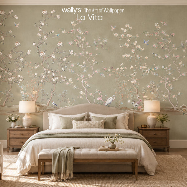 A styled bedroom featuring a large wall mural with the brand name 'wallys The Art of Wallpaper La Vita' at the top. The wallpaper has a sage green background decorated with delicate pink and white cherry blossom branches, blue birds, and small flowers.