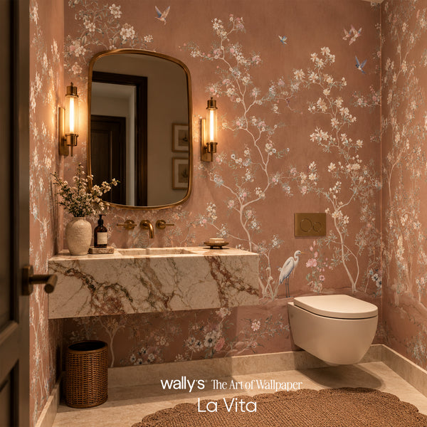 A styled bathroom interior featuring floral wallpaper with white blossoms and blue birds on a dusty pink background, a marble-look floating vanity, wall-mounted fixtures, a toilet, and warm sconces. Text at the bottom reads 'wallys The Art of Wallpaper La Vita'.