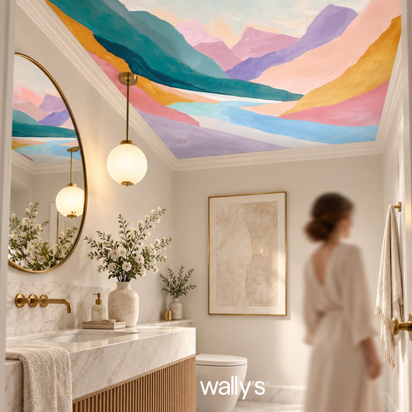 A woman in a white robe stands in a modern bathroom with a marble vanity, round gold-framed mirror, and colorful mountain landscape wall mural. The ceiling also features a matching painted mountain scene.