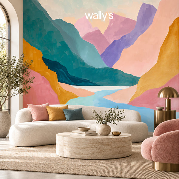 A colorful mountain landscape wall mural in pastel and saturated hues, shown in a styled living room with a cream sofa, round coffee table, pink chair, and gold lamp. The word 'wally's' appears at the top center.