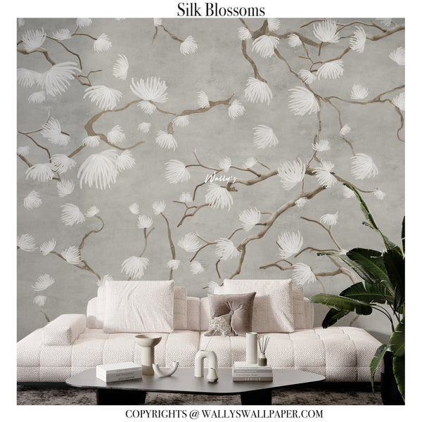 Silk Blossoms wallpaper shown in a styled living room setting, featuring a textured gray background with delicate white and beige cherry blossom branches and flowers. A light beige textured sofa, decorative table, and a green houseplant are placed in the foreground.