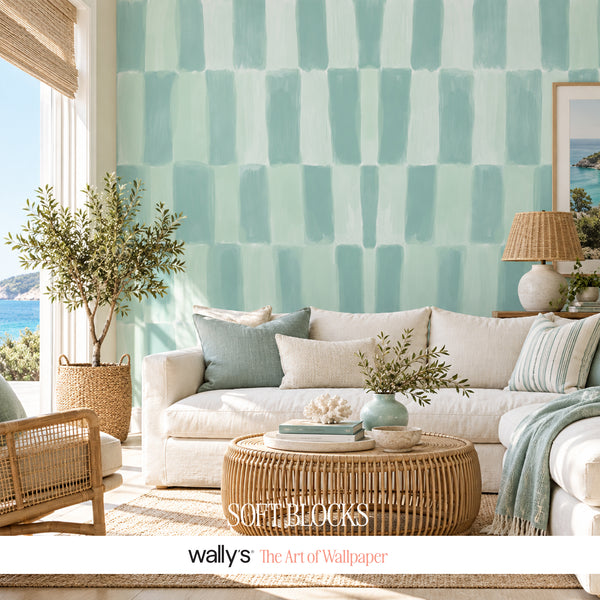 A styled living room featuring a wall with a mint green and aqua soft block wallpaper pattern. The room is furnished with a white sofa, a round woven rattan coffee table, a potted olive tree, and a wicker chair, with a window on the left showing a view of the blue sea.