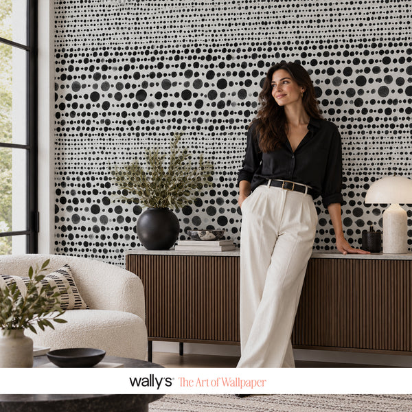 A woman stands in a modern living room wearing a black button-up shirt and white trousers, posing in front of a wall covered in black and white dotted pattern wallpaper. A white armchair, sideboard, and lamps are visible in the room.