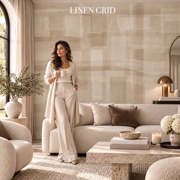 A woman stands in a sunlit living room wearing a matching cream lounge set with a long open-front cardigan. The room features a beige sectional sofa, textured pillows, a stone coffee table, and a large abstract beige wall.